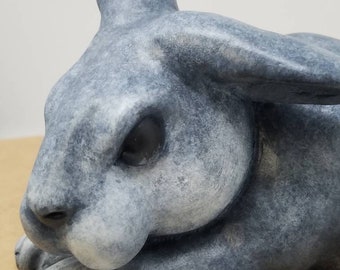 May include: A gray ceramic rabbit figurine with a textured finish. The rabbit is lying down with its head resting on its paws.