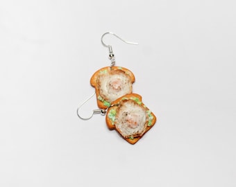 May include: A pair of silver earrings with a fried egg and avocado toast design. The earrings are made of polymer clay and are a fun and quirky accessory.