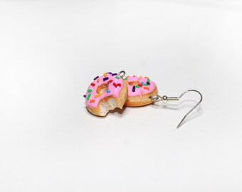 May include: Novelty earrings in the shape of miniature donuts. Each donut features pink icing and colourful sprinkles. One earring has a bite taken out of it. The earrings have silver-coloured fish hook closures.