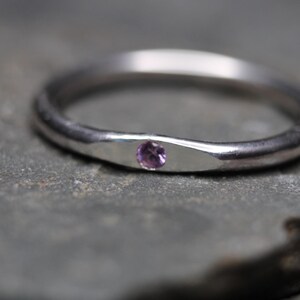 May include: A polished silver-toned metal ring featuring a single, small, round, light purple gemstone set flush into a slightly flattened, tapered section of the band, creating a smooth and continuous circular profile.