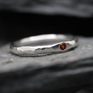 May include: A silver ring with a hammered, textured finish featuring a single, small, round, deep red gemstone set flush into the band. The metal surface has a reflective, polished appearance with irregular indentations across the outer circumference.