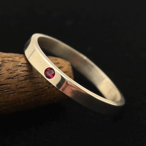 Puede incluir: Un anillo de banda de plata pulida que presenta una única piedra preciosa pequeña, redonda y de color rojo vibrante, engastada al ras en la superficie metálica. El anillo tiene un perfil exterior liso y plano con un ancho constante.