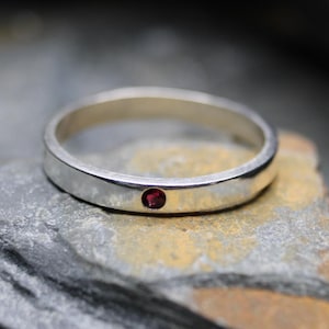 May include: A polished silver band ring featuring a single, small, round, deep red gemstone set flush into the center of the metal surface. The ring has a smooth, flat profile and a reflective finish, showcasing the contrast between the silver and stone.