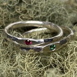 May include: Two silver rings with hammered textures, one with a red gemstone and the other with a green gemstone. The rings are set against a backdrop of light green, textured moss.