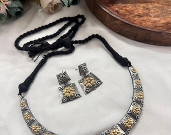 Puede incluir: Un collar plateado y un juego de pendientes a juego con detalles florales dorados. El collar tiene un cordón negro. Los pendientes son cuadrados. Las joyas se exhiben sobre una superficie blanca con flores en el fondo.