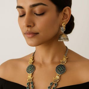 May include: A woman wearing a silver and gold-toned necklace and earrings set. The necklace features green gemstone accents and a floral design. The earrings have a peacock-inspired design. The jewelry is set against a black top.