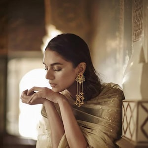 Può includere: Una donna indossa orecchini color oro con un design floreale e fili di perline pendenti. Gli orecchini presentano dettagli rossi e oro. La donna indossa uno scialle color oro.