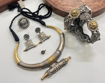 Puede incluir: Un surtido de joyas plateadas y doradas, que incluyen un collar, aretes, un anillo, una pulsera y un colgante. El collar tiene un diseño grueso y curvo con detalles dorados. La pulsera presenta detalles circulares dorados. Las joyas se exhiben sobre una superficie blanca.