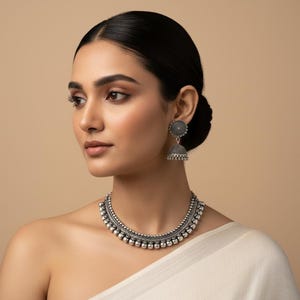 May include: A woman wearing a silver-toned necklace and matching earrings. The necklace features a series of small, round beads. The earrings have a floral design on the top and a bell-shaped bottom. The jewelry is set against a neutral background.