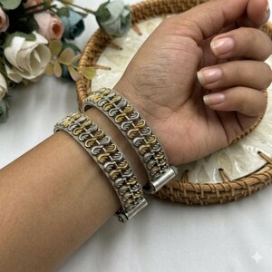 May include: Two silver and gold-toned bangles with a chain-like design are displayed on a wrist. The bangles have a repeating pattern of interlocking links, with alternating silver and gold segments. The clasp is a simple, rectangular design. The background includes a woven basket and flowers.