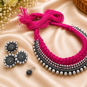 May include: A jewelry set consisting of a braided hot pink thread necklace with silver-toned metal beads, two matching circular stud earrings with intricate embossed patterns, and a single small matching nose ring with a silver-toned finish.