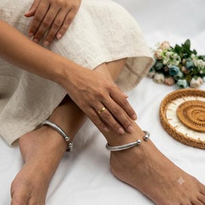 Könnte beinhalten: Ein Paar silberfarbene Fußkettchen mit einem strukturierten, geflochtenen Band und glatten, abgerundeten Kugelenden an beiden Seiten. Die Fußkettchen sind offen gestaltet, was eine flexible Anpassung ermöglicht, und haben eine polierte Oberfläche.