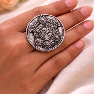 May include: A large, silver ring with an engraved face design. The ring is round and features intricate details, including floral and geometric patterns. The ring is worn on a finger, showcasing its size and design. The ring's diameter is approximately 3.8 cm.