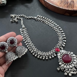 May include: A silver-coloured necklace and earring set featuring a large, round, red gemstone pendant. The necklace and earrings have an ornate, detailed design with smaller red gemstones and a chain.