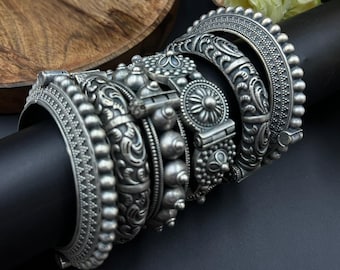 May include: A collection of silver-toned bangles with intricate designs. The bangles feature various patterns, including floral motifs, beaded textures, and detailed scrollwork. The jewelry is displayed on a black cylindrical stand, showcasing the ornate craftsmanship.