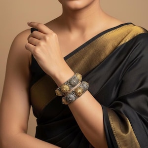May include: Two ornate silver and gold bangles are worn on a wrist. The bangles have a detailed floral design. The person is wearing a black garment with gold trim. The background is a solid tan colour.