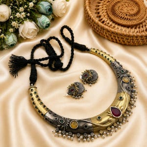 Puede incluir: Un conjunto de collar y pendientes en tonos dorados y plateados. El collar presenta un diseño curvo con piedras preciosas verdes, amarillas y rojas, y pequeñas perlas blancas. Los pendientes tienen un diseño similar con piedras rojas.