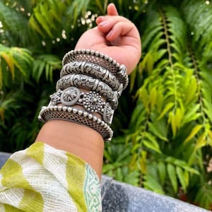 Op de afbeelding: Een verzameling zilverkleurige armbanden met ingewikkelde bloemen- en geometrische ontwerpen. De armbanden variëren in breedte en stijl, sommige hebben kralenranden en gedetailleerde patronen. De sieraden zijn op een pols gestapeld en tonen het vakmanschap en de textuur.