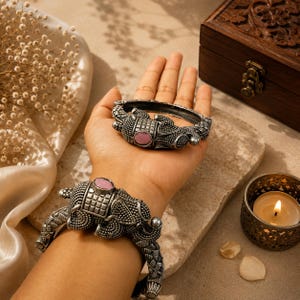 May include: Two silver-toned elephant-shaped bangles with pink center stones. One bangle is worn on a wrist, the other is held in a hand. The bangles have detailed patterns and textures. A lit candle and a wooden box are in the background.