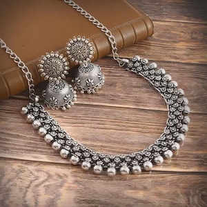 May include: Silver-coloured necklace and earring set. The necklace has a beaded design with a matching chain. The earrings are bell-shaped with floral detailing. The jewellery set is displayed on a wooden surface.