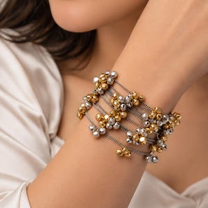 以下が含まれることがあります： ゴールドとシルバーの交互のトーンで、多数の小さな丸い金属製の鈴があしらわれた多連ブレスレット。鈴は、手首に重なり合うように巻き付く、繊細で細いシルバーカラーのチェーンに取り付けられています。