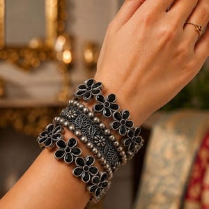 May include: A multi-layered bracelet set featuring two bands with black flower-shaped charms and a central band with intricate filigree leaf patterns and small silver beads. The metal has an oxidised silver finish, creating a dark, textured look.