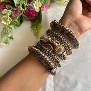 May include: A selection of silver and gold-coloured bangles are displayed on a wrist. The bangles showcase detailed designs, including beaded and embossed patterns. Some bangles have ornate, detailed embellishments. The jewellery is set against a soft, neutral backdrop, with floral accents.