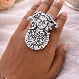 Puede incluir: Un anillo plateado grande con un diseño detallado de rostro. El anillo presenta un rostro ornamentado y estilizado con detalles intrincados. El anillo se lleva en una mano, mostrando el diseño. El anillo es una pieza de joyería llamativa.