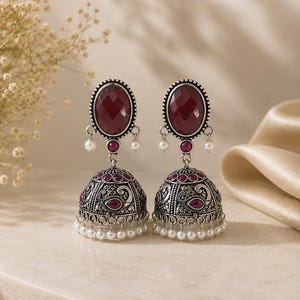 May include: A pair of oxidized silver jhumka earrings featuring large oval faceted deep red stones at the top, smaller round red stones, and bell-shaped drops with intricate embossed patterns, finished with dangling white seed pearls.