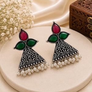 May include: A pair of oxidized silver-tone earrings featuring a triangular base with intricate floral engravings. Each earring is adorned with one teardrop-shaped pink stone, two green teardrop stones, and a row of small white pearl-like beads.