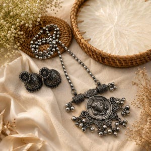 May include: A silver-toned jewelry set featuring a necklace and matching earrings. The necklace has a pendant with a detailed face design and dangling charms. The earrings are round with intricate patterns. The set is displayed on a cream-colored fabric.