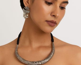 Puede incluir: Collar plateado ornamentado y pendientes a juego. El collar presenta una banda curva y grabada con un colgante triangular adornado con intrincados patrones y pequeñas cuentas blancas. Los pendientes tienen un diseño similar.