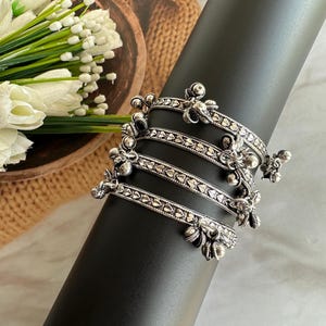 Op de afbeelding: Zilverkleurige armbanden met ingewikkelde bloemen- en geometrische ontwerpen worden getoond op een zwarte cilindervorm. De armbanden hebben een gedetailleerd patroon en zijn op elkaar gestapeld. Witte bloemen en groen zijn op de achtergrond.