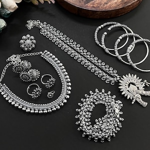 Könnte beinhalten: Eine Sammlung von silberfarbenem Schmuck, darunter eine Halskette, Ohrringe, Ringe, Armbänder und ein Anhänger. Die Stücke weisen aufwendige, detaillierte Designs mit einem Vintage-Look auf. Der Schmuck ist auf einer dunklen Oberfläche angeordnet.