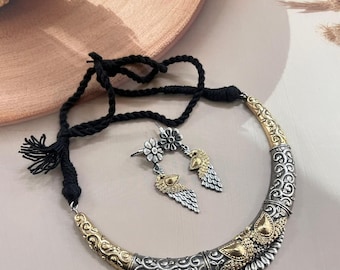 Puede incluir: Un conjunto de collar y pendientes plateados y dorados. El collar tiene un cordón negro y un colgante curvo y ornamentado con detalles dorados. Los pendientes presentan un perno floral y una gota en forma de pluma. Las joyas se exhiben sobre una superficie neutra.