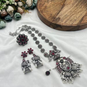 May include: A silver-toned jewelry set featuring a necklace, ring, earrings, and a small pendant. The necklace and ring have a floral design with red accents. The earrings feature elephant pendants with dangling pearls. The set is displayed on a white surface.