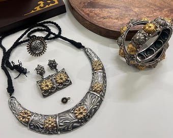 Puede incluir: Un conjunto de joyas plateadas con detalles dorados, que incluye un collar, pendientes, un anillo y una pulsera. El collar tiene forma de media luna con diseños florales. La pulsera y los pendientes tienen patrones florales similares.