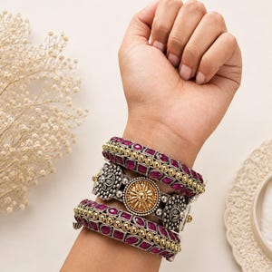 May include: A set of three bangles featuring a central piece with a gold-toned circular floral motif flanked by silver-toned filigree accents, paired with two matching bangles inlaid with deep magenta stones and gold-toned beaded borders.