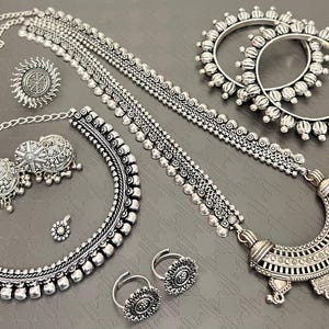 May include: An assortment of silver-toned jewelry pieces, including a necklace, earrings, rings, a nose ring, and two bangles. The jewelry features intricate designs and textures, with a vintage or antique aesthetic. The pieces are arranged on a gray surface.