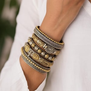 May include: An array of gold and silver bangles, featuring detailed floral and geometric designs, are stacked on a wrist. The bracelets have beaded and engraved elements. The bangles create a layered effect, set against a white backdrop.