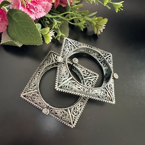 Peut inclure: Deux bracelets carrés en argent avec des motifs de filigrane complexes. Les bracelets sont ouverts et ont une fermeture à charnière. Le fond est noir avec des fleurs roses et vertes.