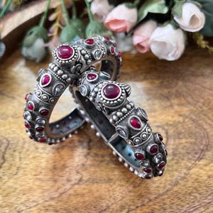 Puede incluir: Dos pulseras de plata ornamentadas con detalles de piedras preciosas rojas. Las pulseras presentan intrincados detalles y están adornadas con múltiples piedras rojas y piedras claras más pequeñas. Las joyas se exhiben sobre una superficie de madera, con flores en el fondo.