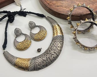 Puede incluir: Un conjunto de joyas plateadas y doradas con un collar, pendientes, un anillo y dos brazaletes. El collar y los pendientes tienen forma de media luna con intrincados diseños florales. Los brazaletes tienen un diseño puntiagudo.