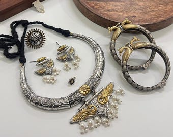 Puede incluir: Un surtido de joyas plateadas y doradas, que incluyen un collar, aretes, un anillo y brazaletes. El collar tiene un cordón negro y un colgante curvo con dibujos. Los aretes y el colgante presentan detalles dorados y perlas.