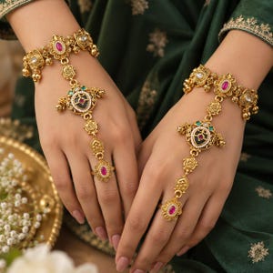 May include: A pair of gold-toned hand harnesses featuring intricate filigree metalwork, small white beads, and central oval-shaped pink stones. Each piece connects a wrist bracelet to a finger ring with a decorative chain and a central medallion.