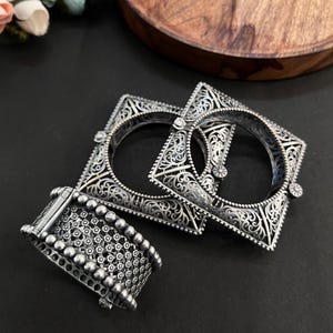 Può includere: Tre braccialetti d'argento con intricati disegni a filigrana e floreali. Due sono quadrati e uno è un bracciale più largo con un motivo a pois. I gioielli sono esposti su una superficie scura.