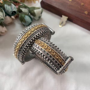 May include: Two silver bangles with gold accents. The bangles feature a detailed design with small, raised silver beads and gold circular patterns. The jewelry is displayed on a white surface.