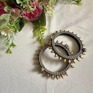 May include: Two silver and gold-toned metal bangles with a spiked design. The bangles are circular and feature alternating silver and gold spikes around the outer edge. The background is a white fabric with pink and white flowers.
