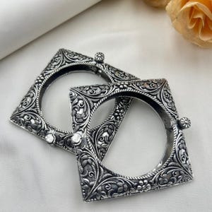 Puede incluir: Dos pulseras cuadradas plateadas con intrincados diseños florales y de hojas. Las pulseras tienen un aspecto vintage, con tallas detalladas y un acabado ligeramente oxidado. Se observan pequeños adornos redondos en los laterales.