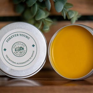 May include: Two open and closed silver tins of skincare balm. The closed tin has a label that reads "FOREVER YOUNG" and "OLD VILLAGE APOTHECARY". The open tin reveals a bright yellow balm. The text on the label also reads "THE NATURAL ALTERNATIVE TO BOTOX".